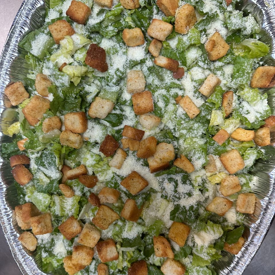 Caesar salad with green lettuce, croutons, and grated cheese in a foil container.